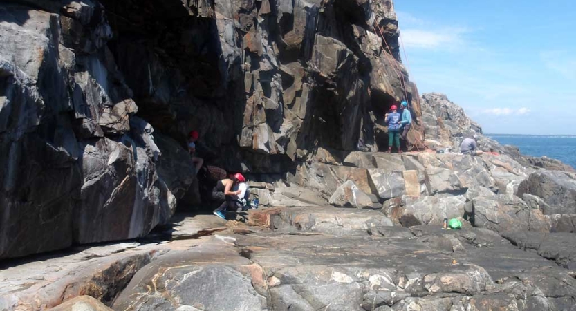 A group of people stand on a rocky shore in safety gear, rock climbing